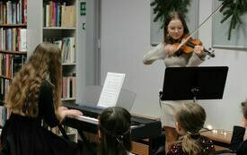 Młoda dziewczyna z długimi włosami i czarnej sukience gra na klawiszach. Obok dziewczyna gra na skrzypcach.