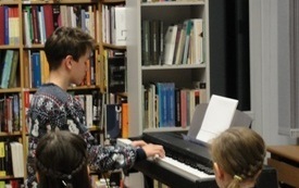 Młoda chłopak w świątecznym swetrze gra na klawiszach. Obok dwie dziewczynki w odświętnych strojach.