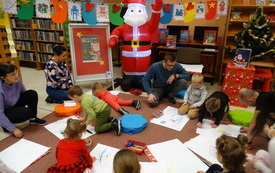 Grupa dzieci wraz z opiekunami wykonuje prace plastyczne. Dzieci siedzą na dywanie. Obok postać Świętego Mikołaja, na lince zwieszone obrazki i skarpety świąteczne.