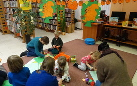 Grupa dzieci podczas wykonywania prac plastycznych w bibliotece. W tle regały biblioteczne. Między regałami zawieszony na lince obrazki przedstawiające wiewi&oacute;rki. 
