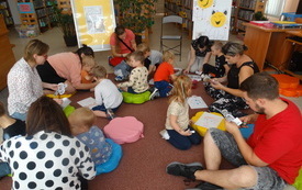 Na dywanie w bibliotece grupa dzieci wraz z opiekunami. W tle regały biblioteczne.