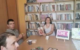 Kilka osób dyskutuje przy stole, kobieta trzyma w ręku książkę.