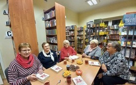 Grupa kobiet siedzi przy stole, w tle kilka regał&oacute;w z książkami. 