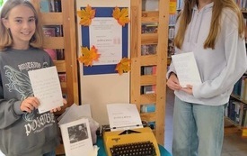 Dwie dziewczyny stoją przy stoliku na kt&oacute;rym stoi maszyna do pisania i książka. Dziewczyny trzymają rękach zapisane kartki papieru. W tle regały biblioteczne.