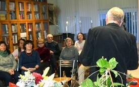 Mężczyzna stoi tyłem, grupa ludzi siedzi na krzesłach i słucha. W tle gabloty z książkami.