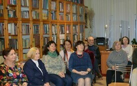 Grupa ludzi siedzi na krzesłach i słucha, w tle gabloty z książkami.