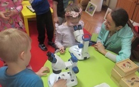 Zielony stoliczek, na nim dwa mikroskopy, lupa, pudełka drewniane. Dziewczynka i chłopiec, pod opieką kobiety oglądają owady pod mikroskopem. 