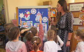 Bibliotekarka stoi wraz z dziećmi przy tablicy, na której są papierowe bałwany.