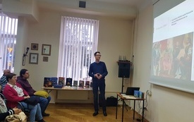 Mężczyzna w okularach zwrócony twarzą do prezentacji na ścianie. Po lewej pierwszy rząd publiczności.