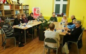 Ludzie siedzą przy stole i rozmawiają, w tle okno.