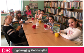 Grupa dziewczynek siedzi przy stole, prezentują książki, kobieta na końcu stołu trzyma zakładki Dyskusyjnych Klub&oacute;w Książki.