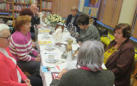 Grupa ludzi siedzi przy stole, na kt&oacute;rym jest wazon z kwiatami, książki, filiżanki, talerze. 