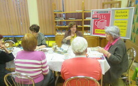 Kobiety siedzą przy stole i rozmawiają, w tle plakat Dyskusyjnych Klub&oacute;w Książki. 