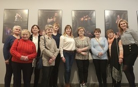 Grupa kobiet stoi obok siebie, obok nich bibliotekarka. W tle plakaty teatralne w ramach.