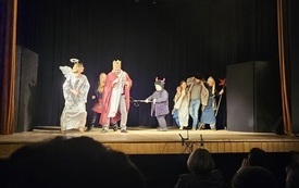 Grupa ludzi na scenie teatralnej przebrana w kostiumy- anioła, diabla, króla.