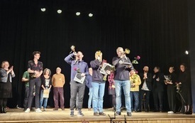 Scena teatralna, trzech mężczyzn stoi na środku i gra na trąbkach, w rękach trzymają róże i papierowe