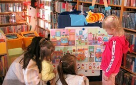 Przy tablicy z kolorowymi obrazkami małe dziewczynki i kobieta. Wokół regały biblioteczne.