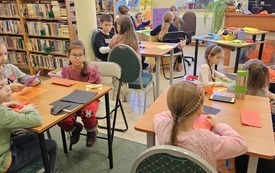 Przy stolikach w bibliotece siedzi grupa dzieci i wykonuje koty metodą origami. 