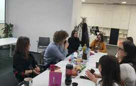 Grupa młodych ludzi siedzi przy stole i rozmawia. Na stole logo Dyskusyjnych Klub&oacute;w Książki. 