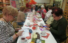 Grupa kobiet siedzi przy stole, ma otwarte książki, zeszyty, w tle balony i plansza dotycząca Walentynek. 
