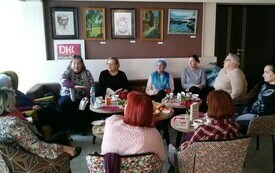 Grupa kobiet w r&oacute;znym wieku siedzi na fotelach i rozmawiają. W tle logo Dyskusyjnych Klub&oacute;w Książki oraz obrazy. 