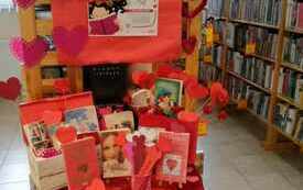Nakryty na czerwono okrągły stolik z książkami, nad nim plakat Zaczytanych walentynek, w tle biblioteczne regały