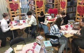 Szybka Randka z Książką - młode osoby siedzą w parach przy stolikach z książkami, w tle udekorowane sercami regały biblioteczne
