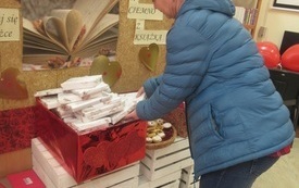 Starsza kobieta pochyla się i wybiera z czerwonego pudełka książkę zapakowaną jak prezent 