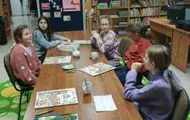 Kilka dzieci siedzi na spotkaniu wok&oacute;ł stołu, na kt&oacute;rym leżą książki. 