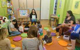 Bibliotekarka siedzi na krzesełku i trzyma książkę, wokół niej siedzą dzieci i rodzice.