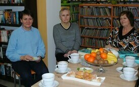Trzy kobiety siedzą przy stole, jedna z nich trzyma w rękach filiżankę. Na drugim planie regały z książkami.