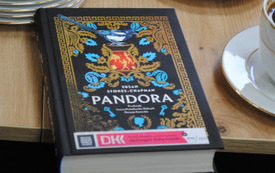 Książka o tytule "Pandora" leży obok talerzyków i filiżanki z kawą.