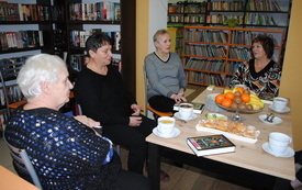Cztery starsze kobiety siedzą przy stole, w tle regały z książkami.