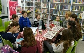Kobieta wraz z dziećmi siedzi przy stole, dzieci coś opowiadają, kobieta się uśmiecha.