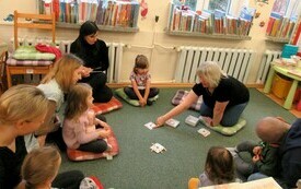 Książki stoja na parapetach. Dzieci i rodzice siedzą na poduszkach. Bibliotekarka prowadzi grę. 