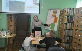 Na ścianie prezentacja dotycząca języka polskiego. Bibliotekarka wskazuje dłonią na tablicę. Dzieci słuchają siedząc przy stolikach. 