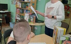 Bibliotekarka w okularach trzyma w ręku kartkę. Przed nią przy stoliku siedzą chłopcy.