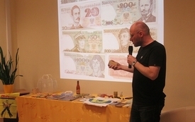 Przy stoliku mężczyzna trzymający w ręku mikrofon. W tle prezentacja banknoty.