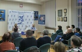 Dwie kobiety o blond włosach siedza na fotelach i rozmawiają. Widownia wsłuchuje się w rozmowę.