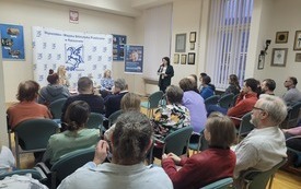 Sala wypełniona widownią. Pośrodku trzy kobiety. Jedna z nich przemawia do mikrofonu.