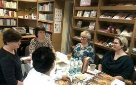 Pięć kobiet siedzi przy stole, rozmawia. W tle regały z książkami, gazetami. 