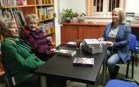 Trzy kobiety siedzą przy stole, rozmawiają. Po lewej stronie regał z książkami. 