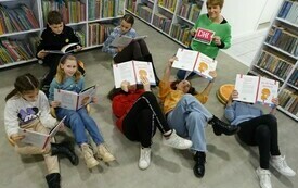 Siedmioro dzieci trzyma książki, troje leży na podłodze, obok nich bibliotekarka trzyma logo DKK. 
