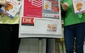 Dziewczynka i bibliotekarka trzymają książki i stoją obok plakatu DKK. 