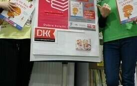 Dziewczynka i bibliotekarka trzymają książki i stoją obok plakatu DKK.