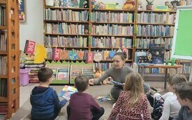 Bibliotekarka i dzieci siedzą na dywanie, w tle loga DKK i regał z książkami. 