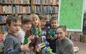 Bibliotekarka wraz z dziećmi trzyma w rękach ufoludki wykonane z papieru. W tle regały z książkami. 