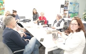Grupa ludzi siedzi przy stole, bibliotekarka pokazuje książkę &quot;Internat&quot; S. Żadana. 