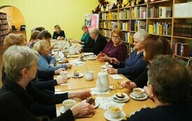 Grupa ludzi w różnym wieku siedzi przy stole i rozmawia, po prawej stronie przy ścianie regały z książkami.