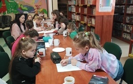 Grupa dzieci siedzi przy stole, na kt&oacute;rym są książki, kubki z logotypem Dyskusyjnych Klub&oacute;w Książki. 
